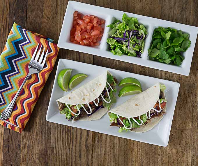 two pork carnitas tacos on a plate with lime wedges, a tray of toppings above, and a fork and napkin on the left