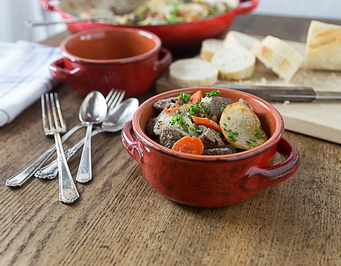 a red bowl of french beef stew next to spoons and forks with bread and a dutch oven in back