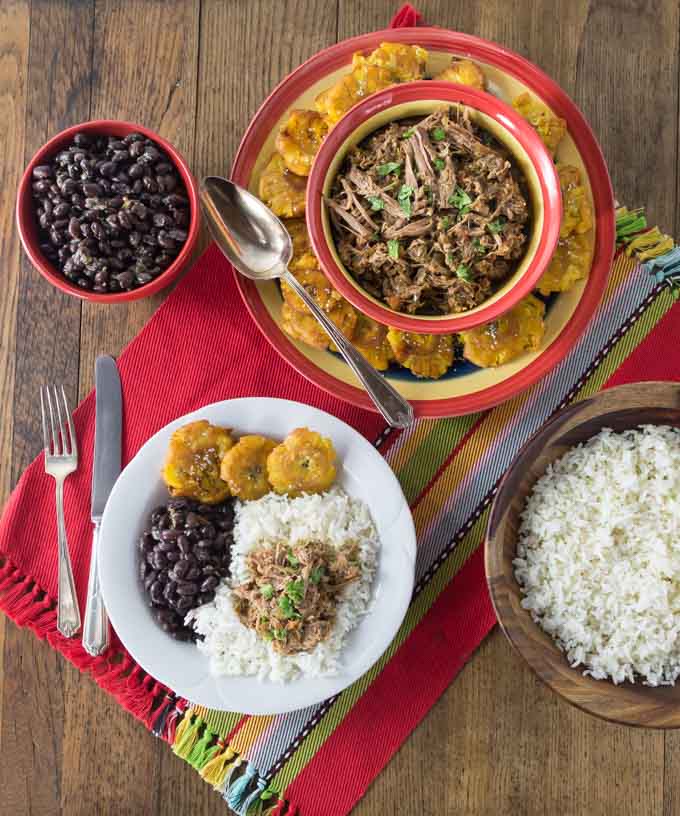 a top down view of a table with ropa vieja, plantains, beans, and rice on a red placemat