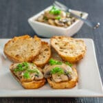 A photo of mushrooms tapas cooked in sherry sitting on baguette with a white plate.